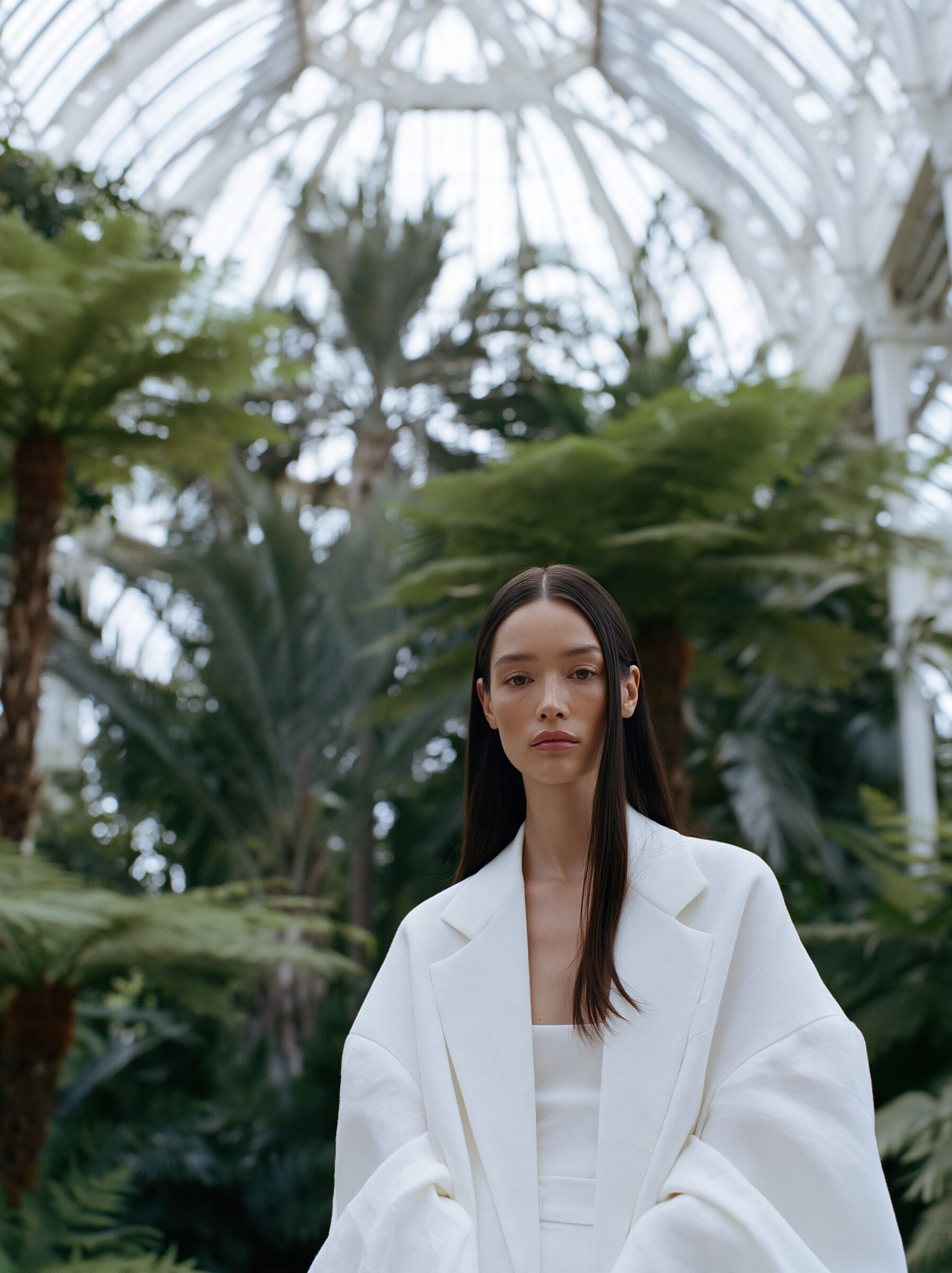 Model in an ivory linen suit standing small among towering palms beneath a curved glass roof