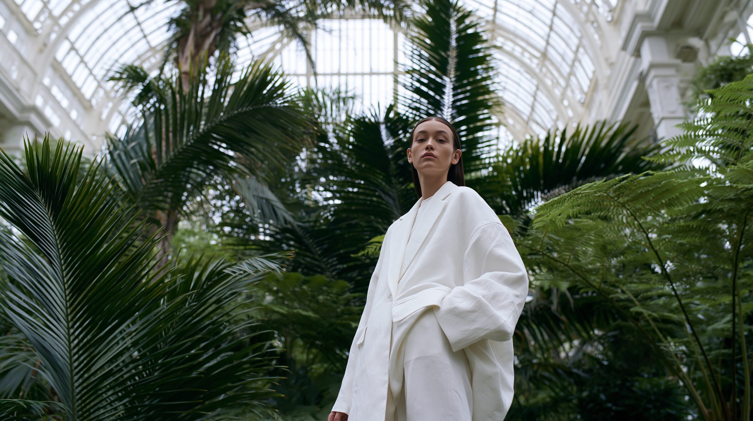 Model in an ivory linen suit standing small among towering palms beneath a curved glass roof