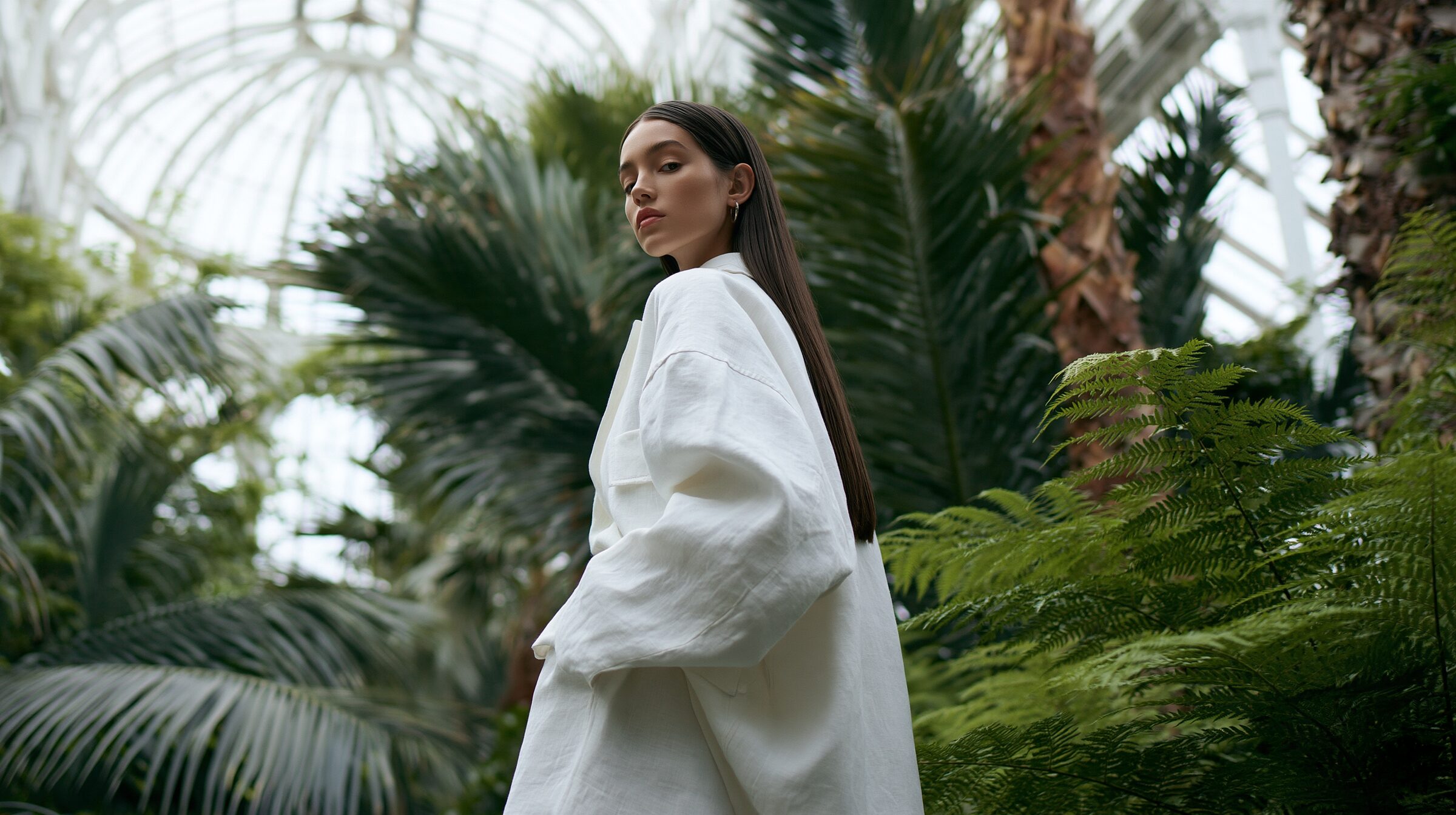 Model in an ivory linen suit standing small among towering palms beneath a curved glass roof