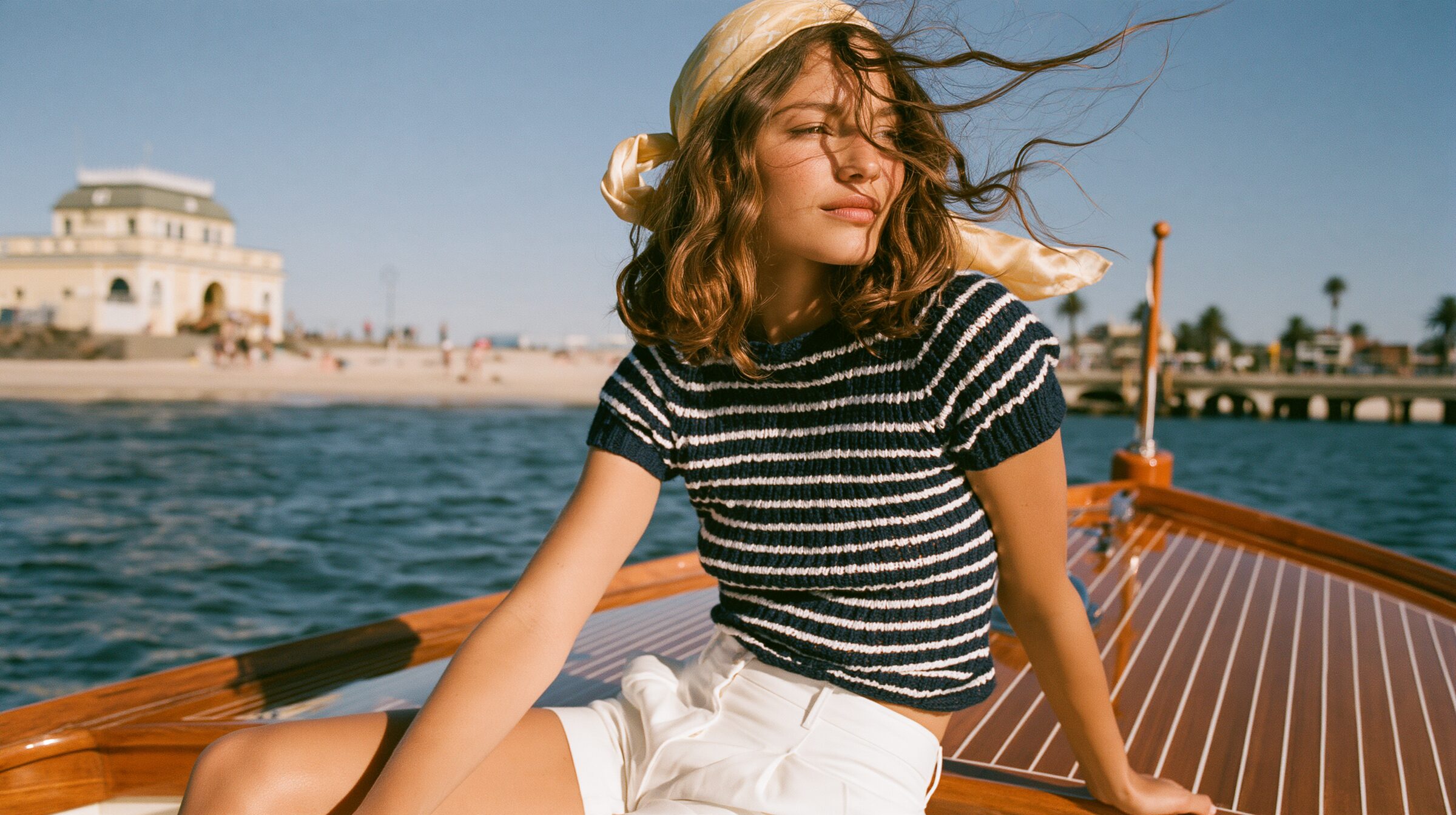 Model seated at the bow of a vintage mahogany speedboat, the Sorrento cliffs behind her